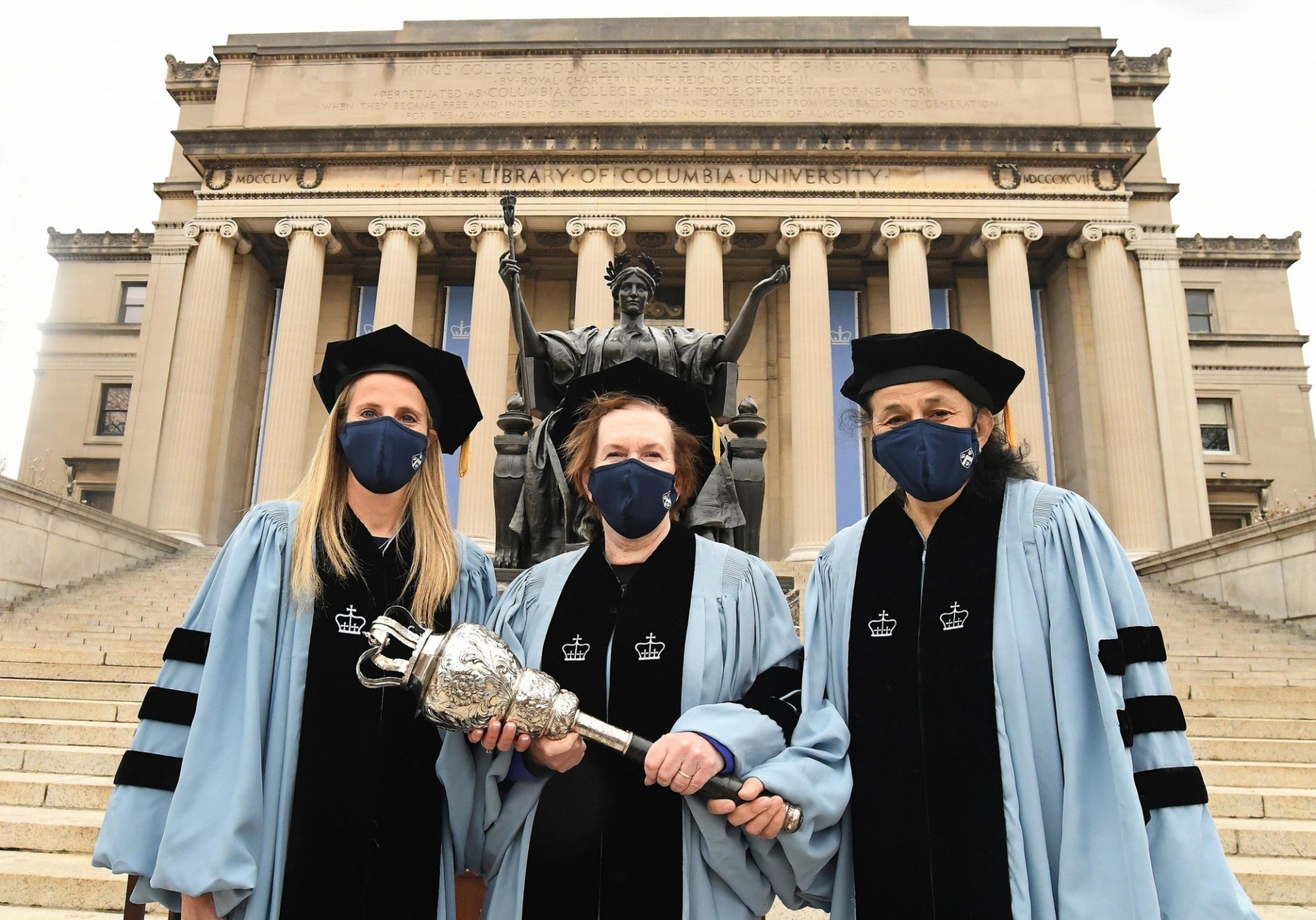 Wearing academic robes, Melanie J. Bernitz, Donna Lynne, and Wafaa El-Sadr hold the University Mace standing in front of Alma Mater.