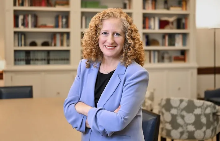 Columbia University President Jennifer L. Mnookin in a light blue jacket with cream colored bookshelves lined with books in the background